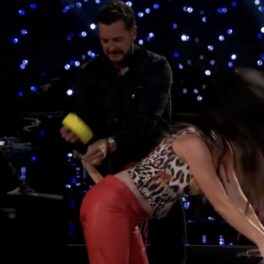 Katy Perry în timp ce se sprijină de o masă, iar un membru din juriul emisiunii la care a participat încearc să-i lipească pantalonii cu bandă adezivă galbenă