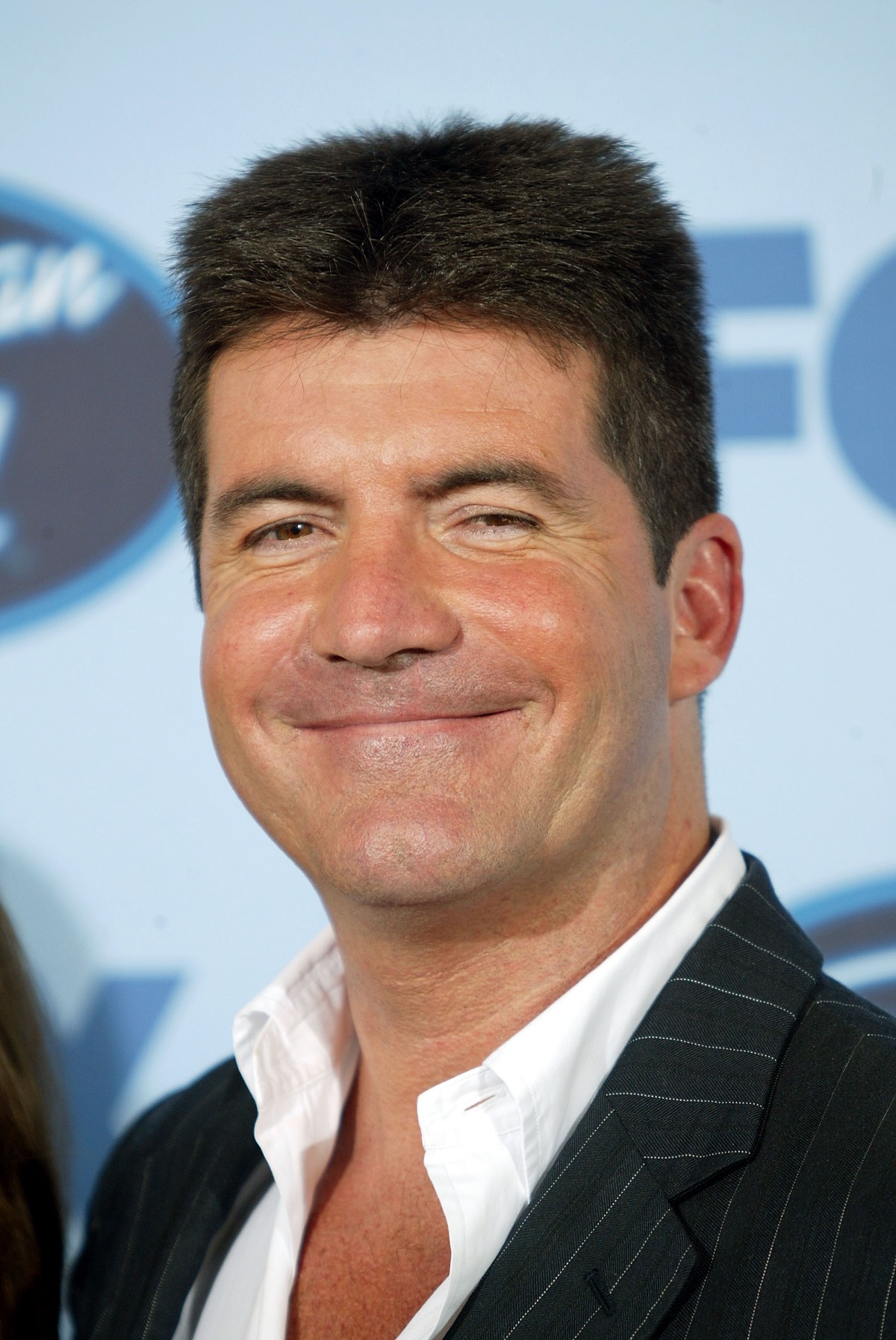 Simon Cowell la costum și cămașă albă, în timp ce zâmbește, înainte de a intra pe platourile de filmare ale American Idol în anul 2005