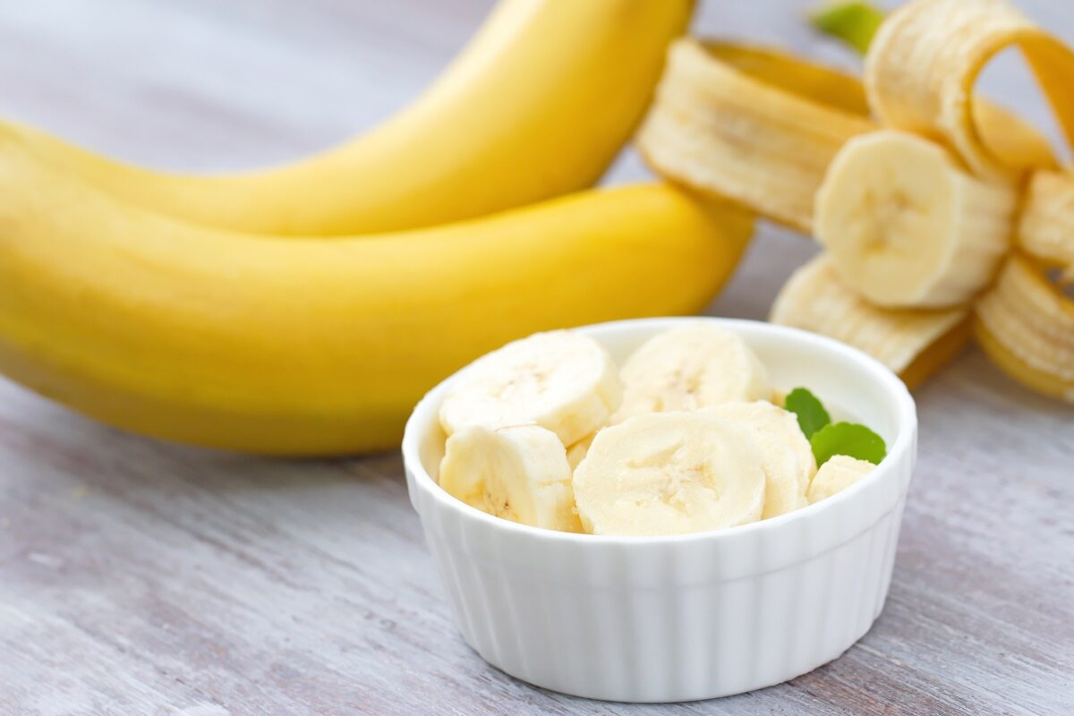 un blat alb e care se află un bol cu felii de banane și două banane coapte pe fundal