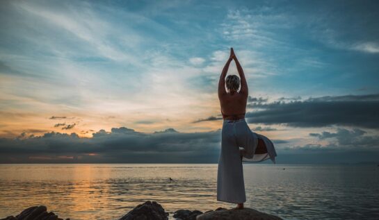 O femeie care stă cu spatele pe o stâncă în timp ce face yoga în fața unei mări pentru a reprezenta una din principalele investiții în sănătate în anul 2022