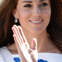 Kate Middleton într-o rochie albă cu imprimeu floral albastru în timp ce face cu mâna publicului și își expune unul din degetele bandajate