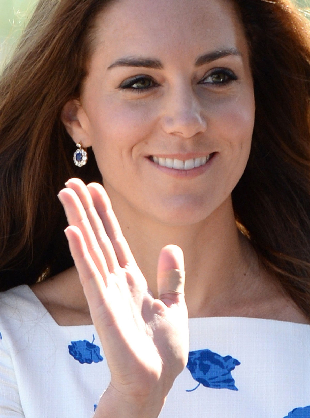Kate Middleton într-o rochie albă cu imprimeu floral albastru în timp ce face cu mâna publicului și își expune unul din degetele bandajate