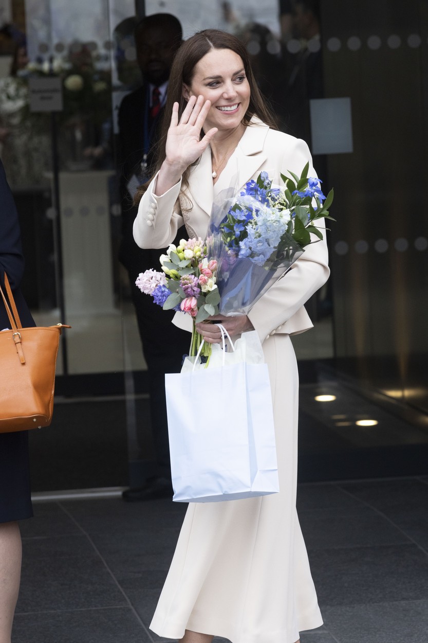 Kate Middleton, îmbrăcată într-un costum deschis la culoare, cu flori în brațe și pantofi cu toc