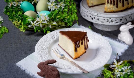 Porție de pască cu mascarpone și ciocolată pe o farfurie albă cu furculiță, alături de un platou cu pasca secționată, un suport floral cu ouă vopsite și un iepuraș din aluat cu cacao