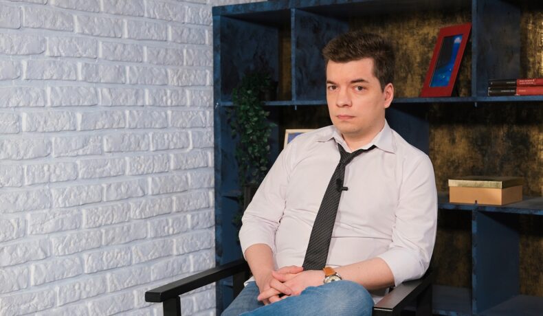 invitatul de la interviul CaTine.ro la masculin, Toma Alexandru, în timp ce oartă o cămașă albă și o cravată neagră și pozează pe platourile de filmare
