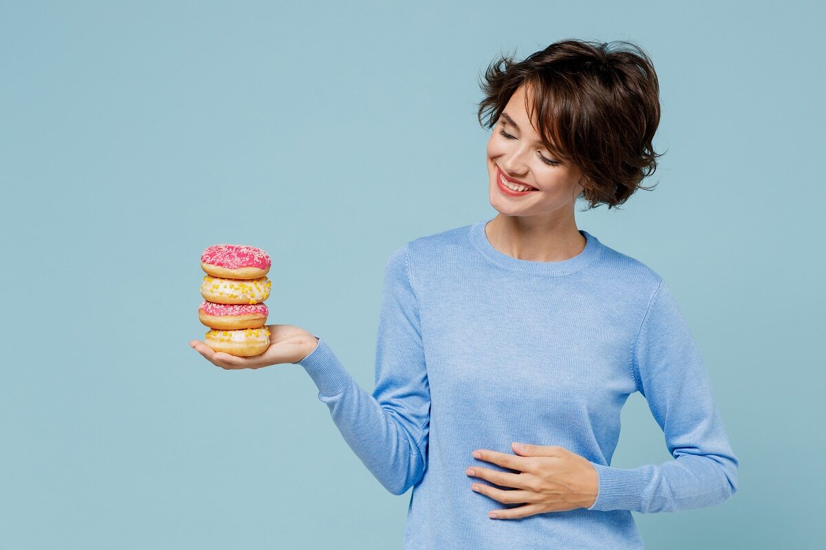 O femeie frumoasă care poartă un tricou albastru în timp ce ține în mână trei gogoși pentru a ilustra unul din principalele obiceiuri dulci care te-ar putea ajuta să reduci grăsimnea abdominală