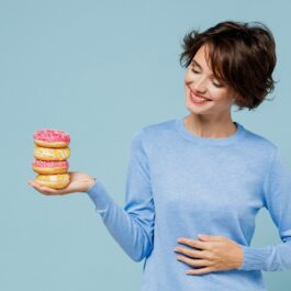 O femeie frumoasă care poartă un tricou albastru în timp ce ține în mână trei gogoși pentru a ilustra unul din principalele obiceiuri dulci care te-ar putea ajuta să reduci grăsimnea abdominală