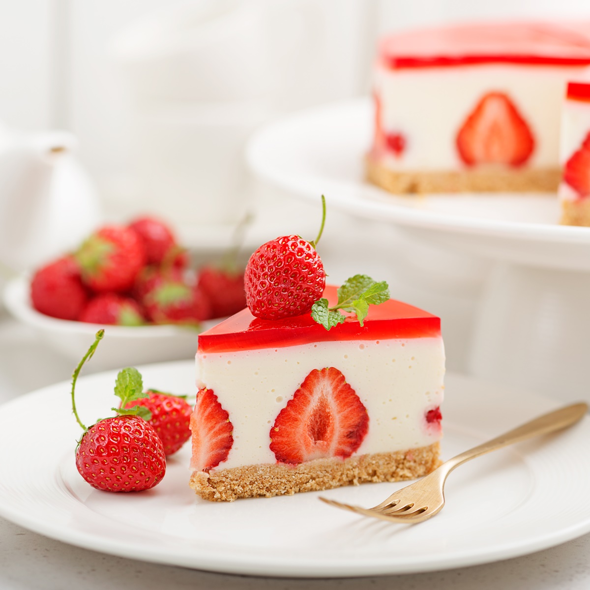 Porție de cheesecake cu jeleu de căpșuni pe o farfurie albă, alături de platoul cu cheesecake și un set de ceai