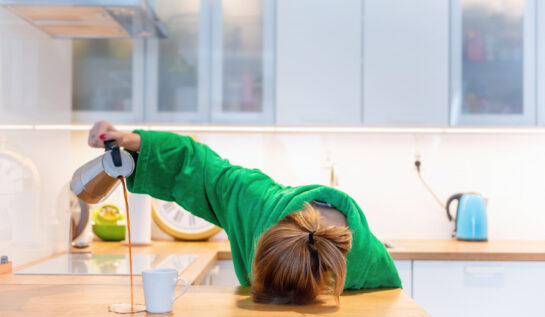 O femeie care este foarte obosită, toarnă cafea în afara cănii și doarme cu capul pe blatul de la bucătărie