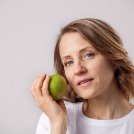 O femeie frumoasă care poartă un tricou alb și ține în mână un măr verde pentru a ilustra acele obiceiuri alimentare care te-ar putea ajuta să reduci grăsimea abdominală și să încetinești procesul de îmbătrânire