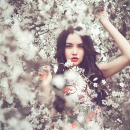 O femeie frumoasă care stă ascunsă între crengile înflorite ale unui copac pentru a ilustra cum să-ți alegi parfumul ideal în funcție de zodie