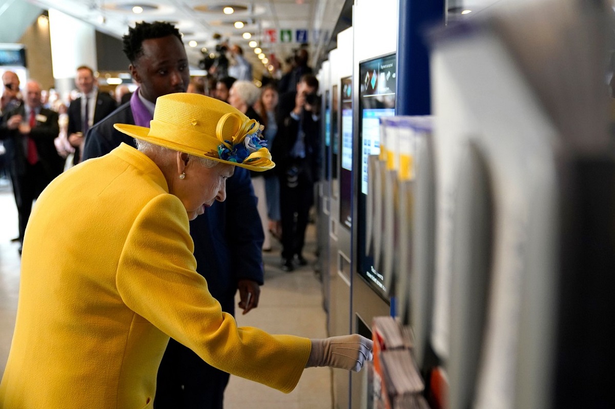 Regina Elisabeta într-un costum galben în timp ce cumpără o cartelă de metrou de la Paddington Station pentru a călătorii cu pe linia de metrou care îi poartă numele, Elizabeth Line