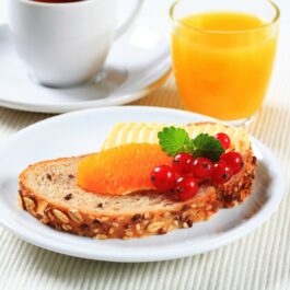 Unt pe o felie de pâine, cu o ceașcă de ceai și un pahar cu suc de portocale alături