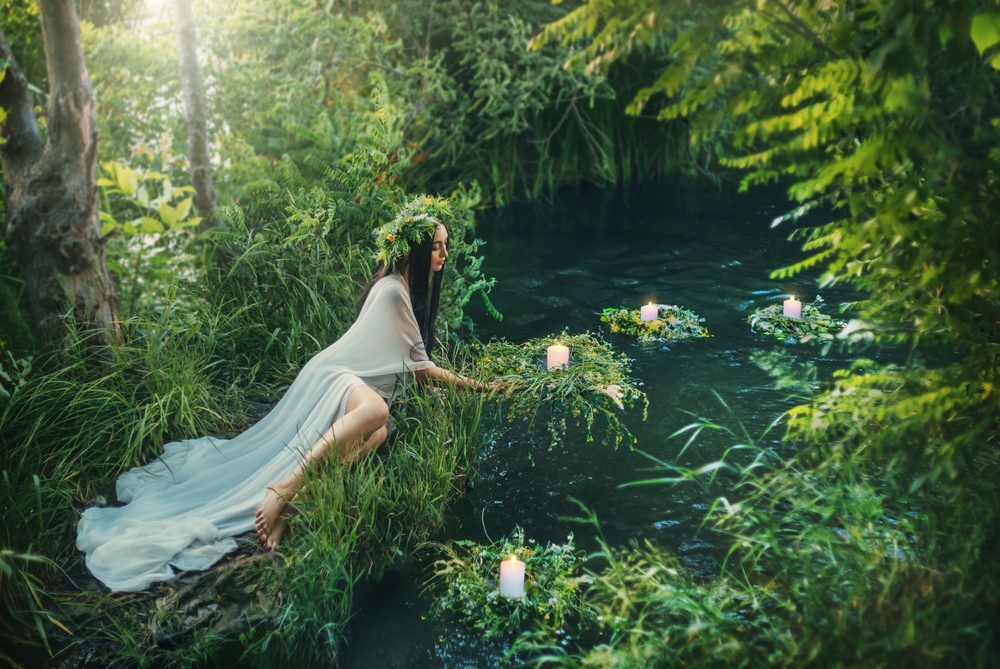 Fată frumoasă îmbrăcată în alb stă la marginea unui râu și poartă o coroniță din flori pe cap