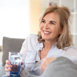 O femeie frumoasă, blondă, care stă pe o canapea și țină în mână un pahar de apă pentru a ilustra acele obiceiuri care grăbesc procesul de îmbătrânire