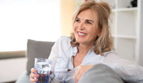 O femeie frumoasă, blondă, care stă pe o canapea și țină în mână un pahar de apă pentru a ilustra acele obiceiuri care grăbesc procesul de îmbătrânire