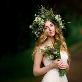 Fată frumoasă îmbrăcată în alb cu o coroană de flori pe cap și cu un buchet de flori în mână