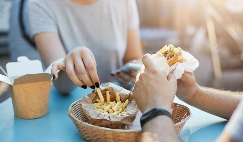 O masă albastră pe care se află un coșuleț cu cartofi prăjiți în timp ce un brăbat și o femei mănâncă din ei pentru a ilustra principalele obiceiuri alimentare care favorizează creșterea nivelului colesterolului
