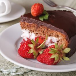 Porție de tort cu mousse de căpșuni și glazură de ciocolată pe o farfurie albă, cu două căpșuni proaspete