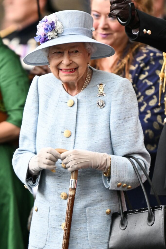 Regina Elisabeta, cu o pălărie pe cap, în baston, într-un costum albastru