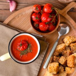 Porție de Supă cremă de roșii coapte, cu usturoi și crutoane aromate în cană de ceramică, alături de un bol cu roșii coapte și o lingură pe o tavă de lemn, rotundă