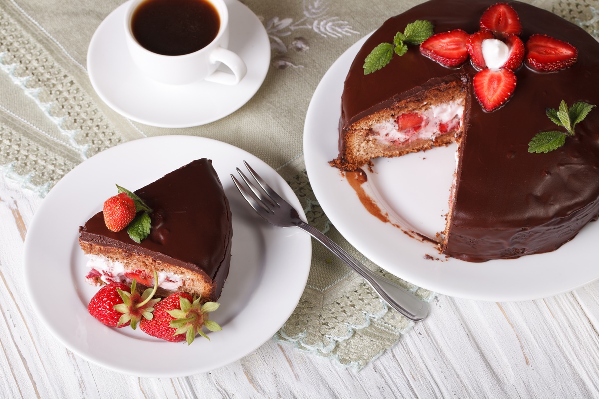 Tort cu mousse de căpșuni și glazură de ciocolată porționat pe o farfurie albă și pe platou, alături de o ceașcă de cafea