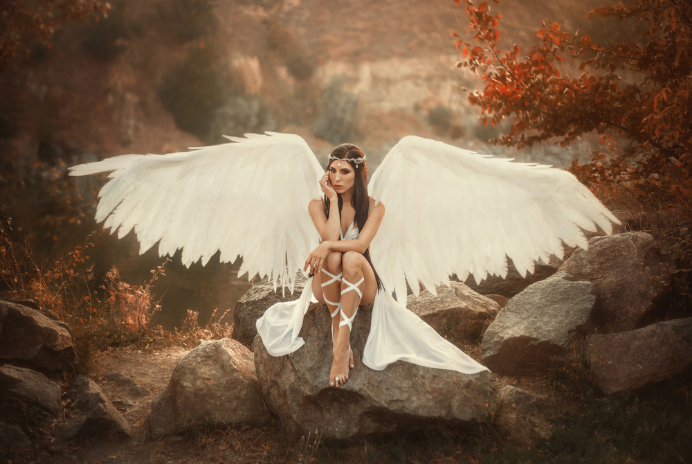 Fată frumoasă cu aripi de înger stă jos și își ține mâinile sprijine de corp
