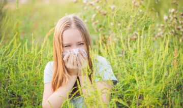 O femeie blondă care stă într-un câmp în timp ce își suflă nasul de alergia la ambrozie