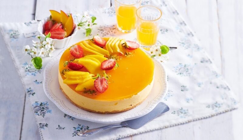Cheesecake fără coacere cu mango pe o farfurie albă, alături de două pahare cu suc și un bol cu felii de fructe