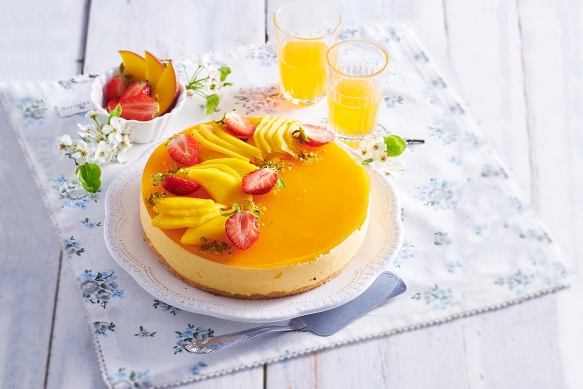 Cheesecake fără coacere cu mango pe o farfurie albă, alături de două pahare cu suc și un bol cu felii de fructe