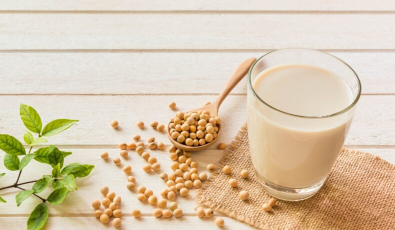 Un blat pe care se află un pahar cu lapte de soia alături de mai multe boabe de soia pentru a demonstra care sunt efectele benefice ale consumului de soia asupra organismului