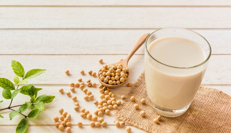 Un blat pe care se află un pahar cu lapte de soia alături de mai multe boabe de soia pentru a demonstra care sunt efectele benefice ale consumului de soia asupra organismului
