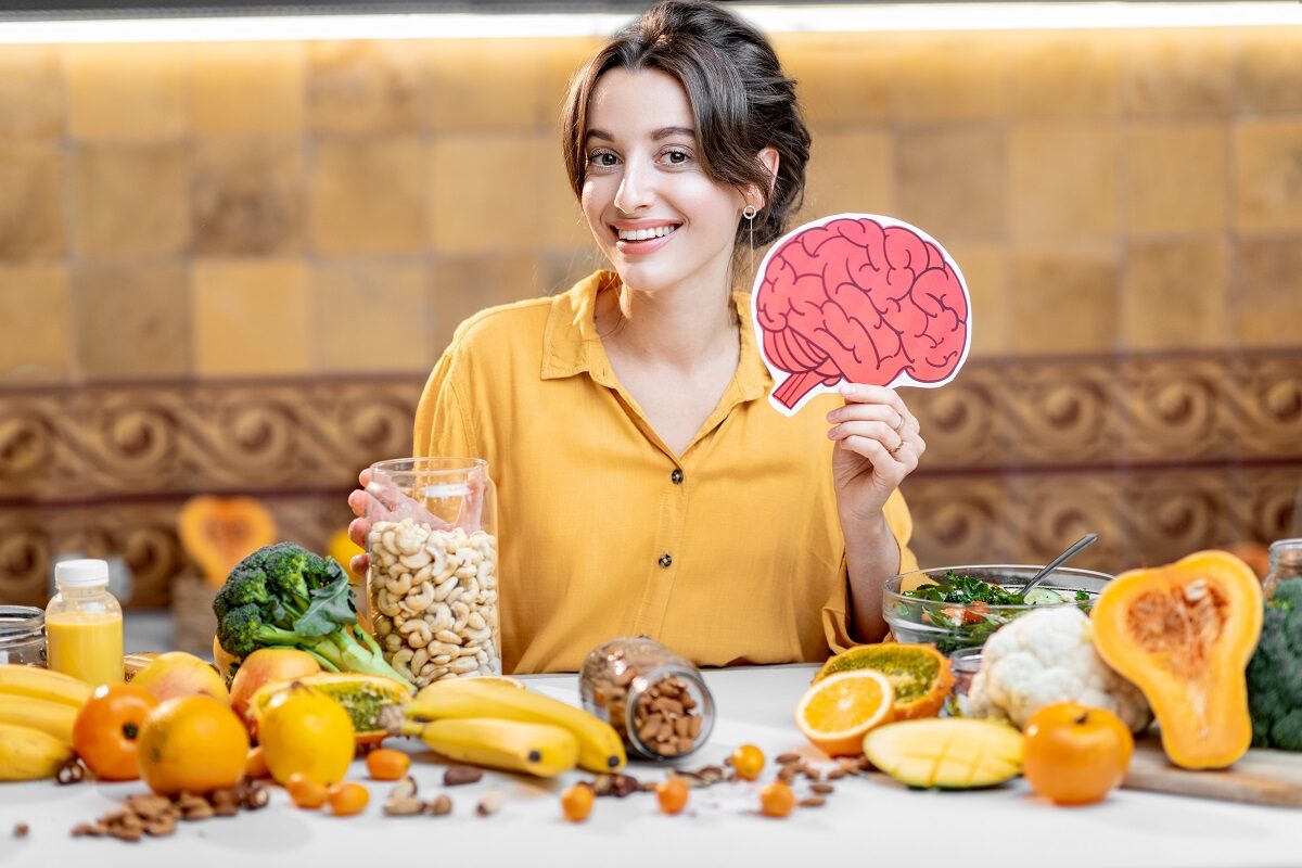 O femeie frumoasă care stă în fața unei mese plină cu fructe și legume în timp ce ține în mână un șablon în formă de creier pentru a ilustra principalele obiceiuri alimentare care îți pot afecta memoria