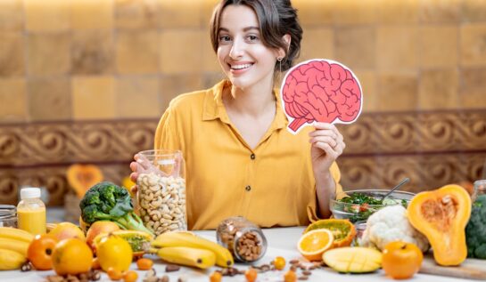 O femeie frumoasă care stă în fața unei mese plină cu fructe și legume în timp ce ține în mână un șablon în formă de creier pentru a ilustra principalele obiceiuri alimentare care îți pot afecta memoria