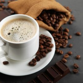 O ceașcă de cafea care se află pe un blat gri alături de o tabletă de ciocolată neagră și câteva boabe de cafea pentru a ilustra câte cești de cafea poți bea zilnic