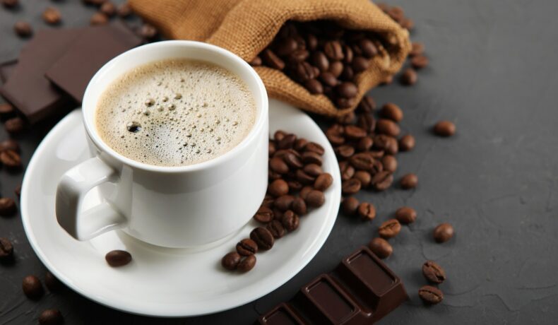 O ceașcă de cafea care se află pe un blat gri alături de o tabletă de ciocolată neagră și câteva boabe de cafea pentru a ilustra câte cești de cafea poți bea zilnic