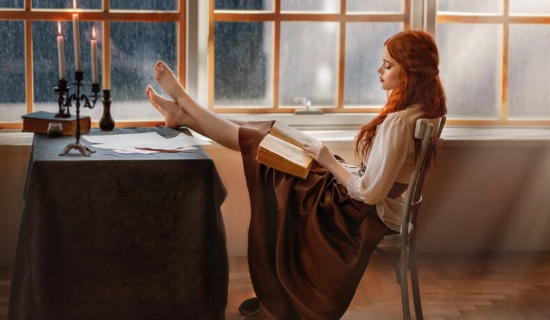 Fată frumoasă cu părul roșcat stă cu picioarele pe masă și citește