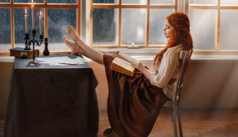 Fată frumoasă cu părul roșcat stă cu picioarele pe masă și citește
