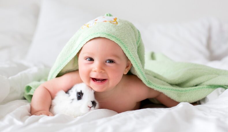 Un bebeluș care stă pe un pat alături de un iepuraș în timp ce zâmbește la cameră