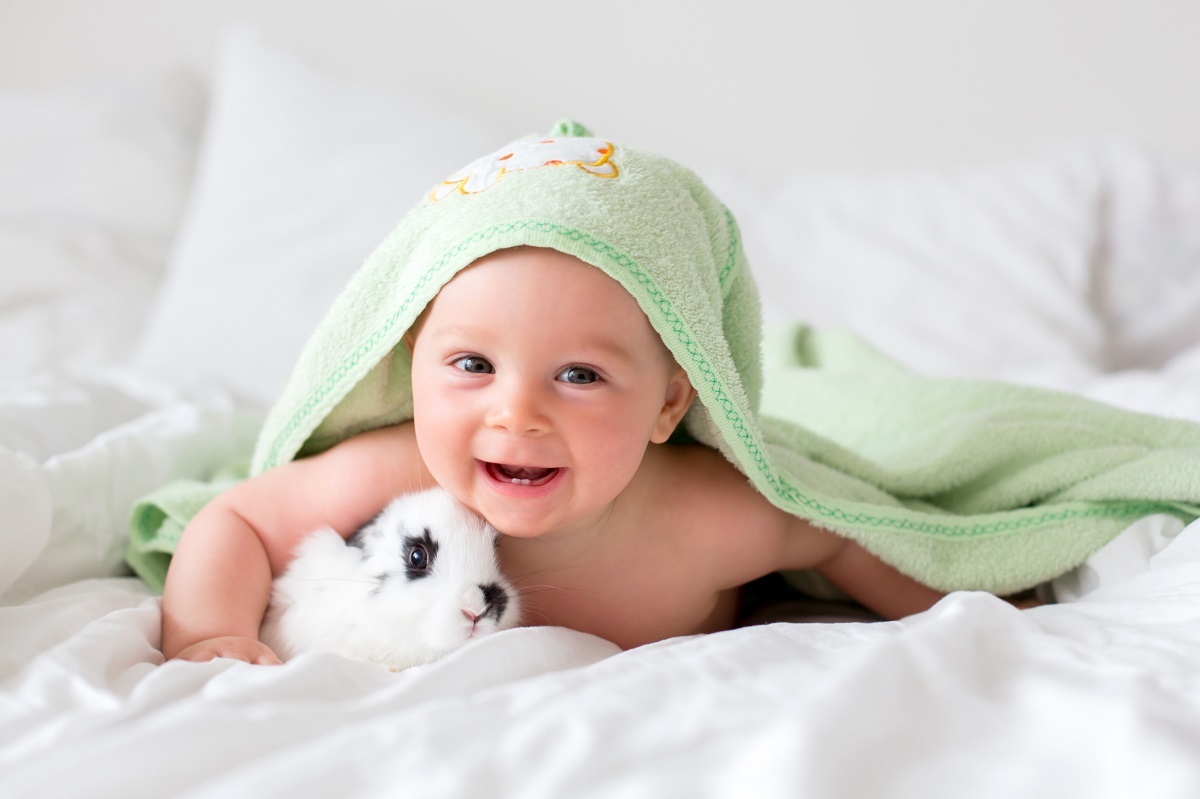 Un bebeluș care stă pe un pat alături de un iepuraș în timp ce zâmbește la cameră