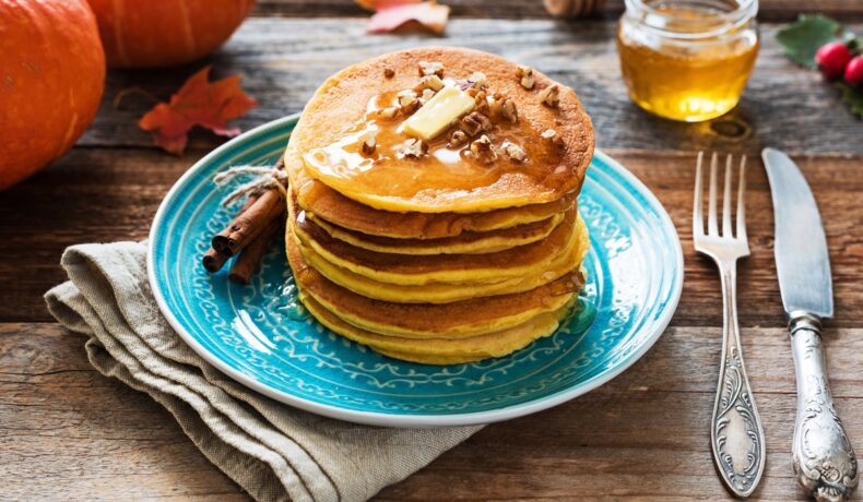 Pancakes cu dovleac plăcintar în aluat așezate pe o farfurie albastră și decorate cu miere, unt și nuci
