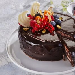 Tort de ciocolată cu glazură oglindă, porționat pe un platou alb, alături de un bol cu fructe de pădure