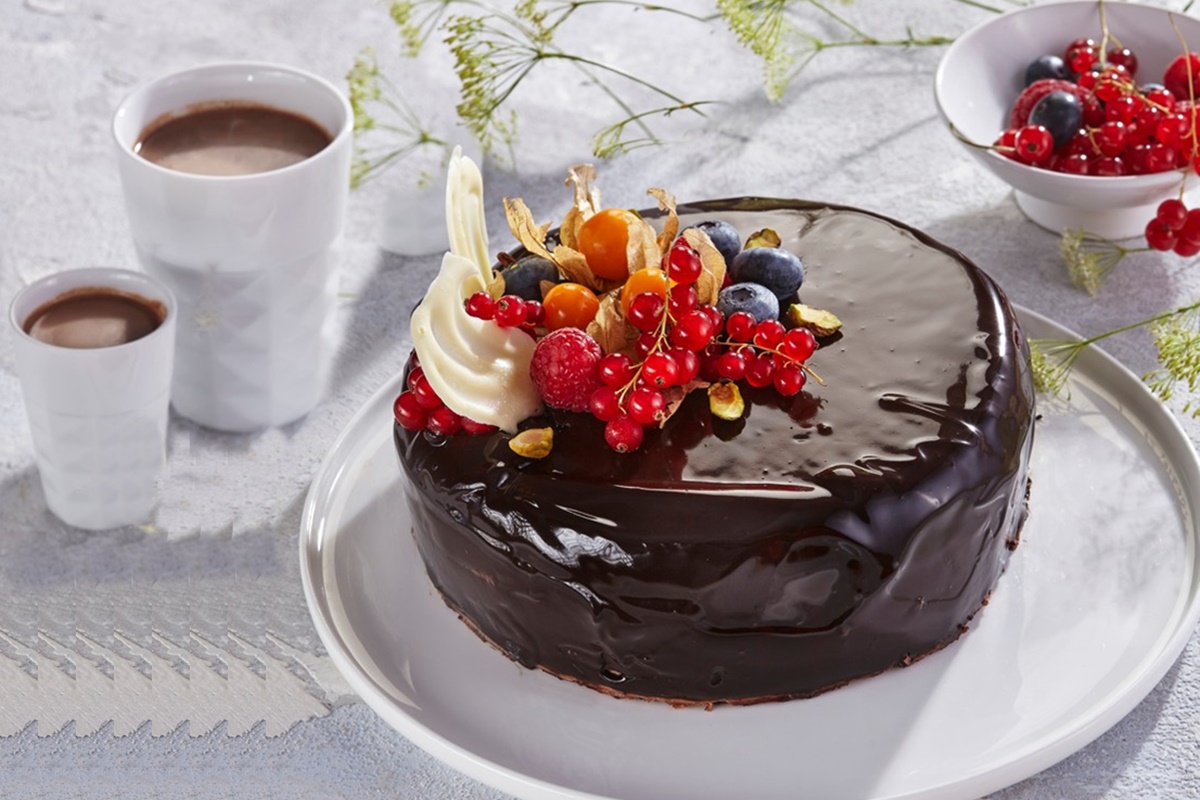 Tort de ciocolată cu glazură oglindă, decorat cu fructe pe un platou alb, alături de două pahare cu cafea și un bol cu fructe