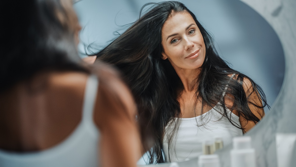 O femeie frumoasă cu părul lung care se uită în oglindă având una din principalele tunsori recomandate pentru femei după vârsta de 40 de ani