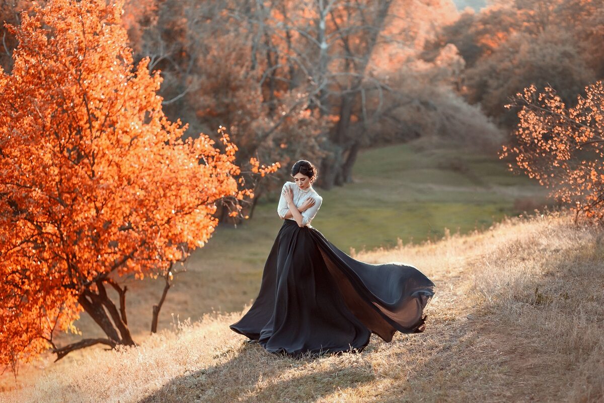 O femeie frumoasă îmbrăcată într-o rochie neagră care se află pe dealuri cu copaci cu frunze ruginii pentru a reprezenta una din cele trei zodii ghinioniste în ziua de 24 octombrie 2022