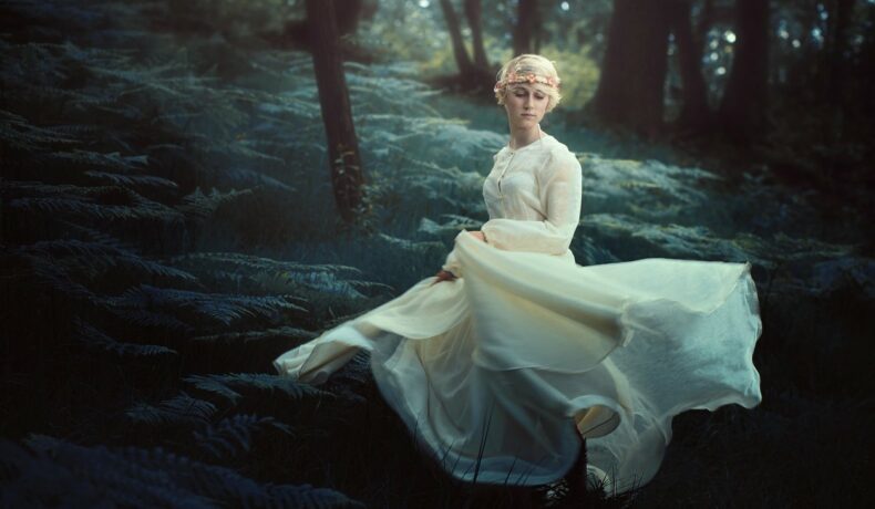Fată frumoasă îmbrăcată într-o rochie lungă, albă, stă în pădure