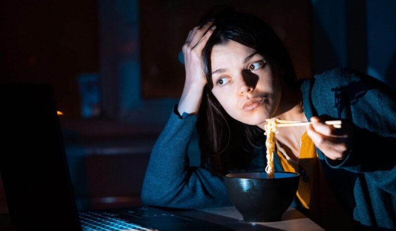 O femeie tânără care mănâncă noaptea în timp ce lucrează la laptop