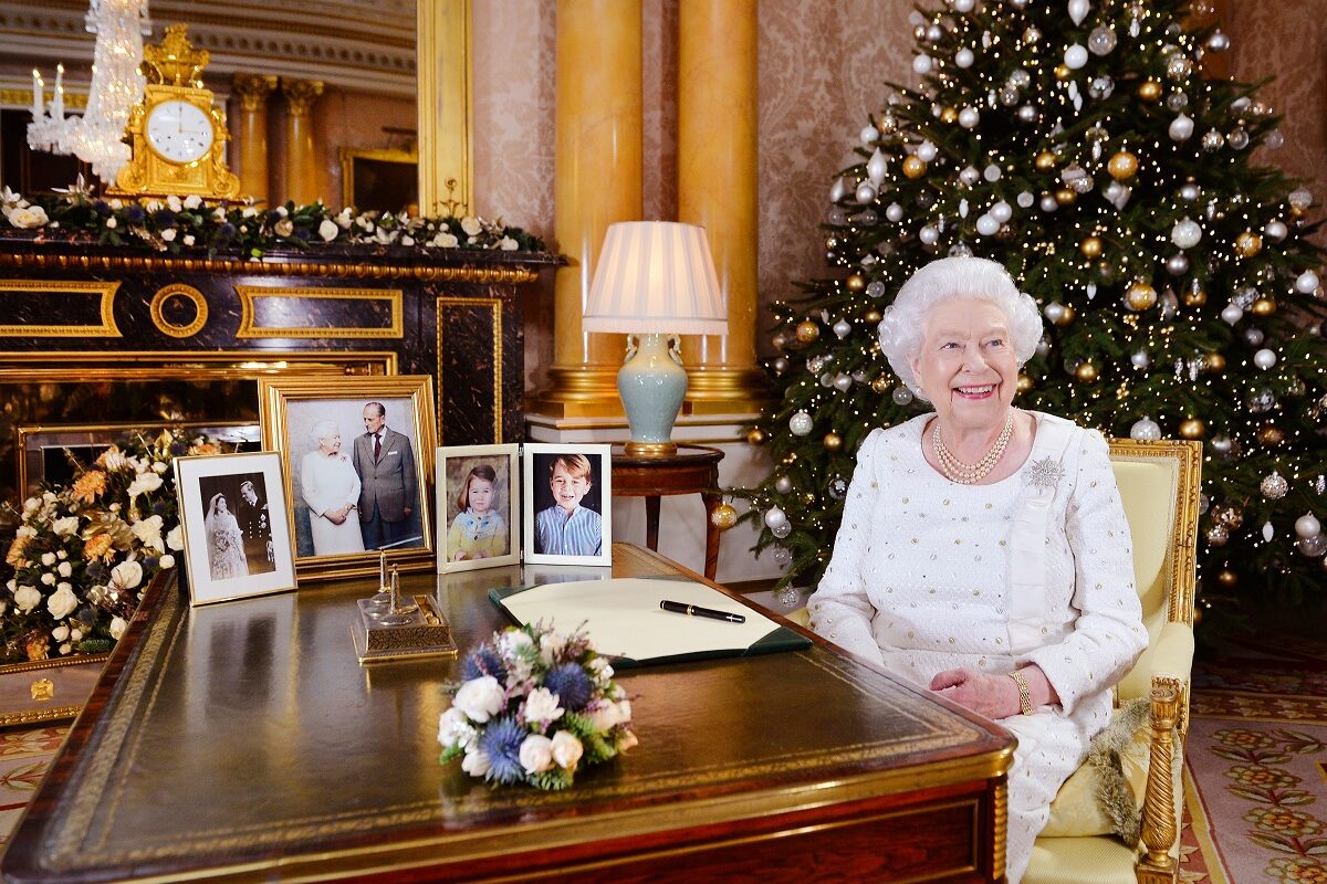 Regina Elisabeta a II-a purtând o rochie albă în timp ce se află la biroul său de la CAstelul Windsor alături de bradul de Crăciun și câteva fotografii ale Familiei Regale Britanice