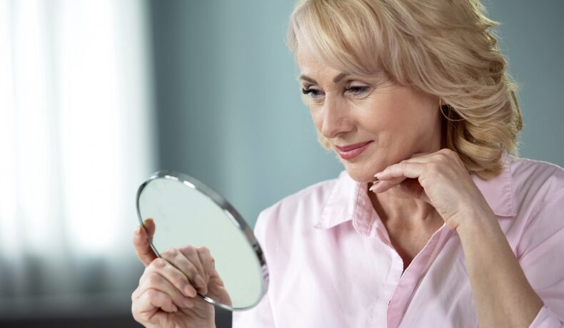 O femeie frumoasă, blondă, care are peste 50 de ani și se privește în oglindă