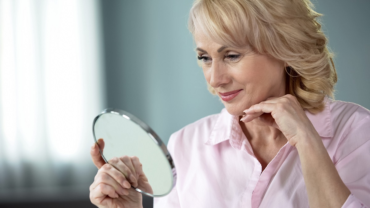 O femeie frumoasă, blondă, care are peste 50 de ani și se privește în oglindă
